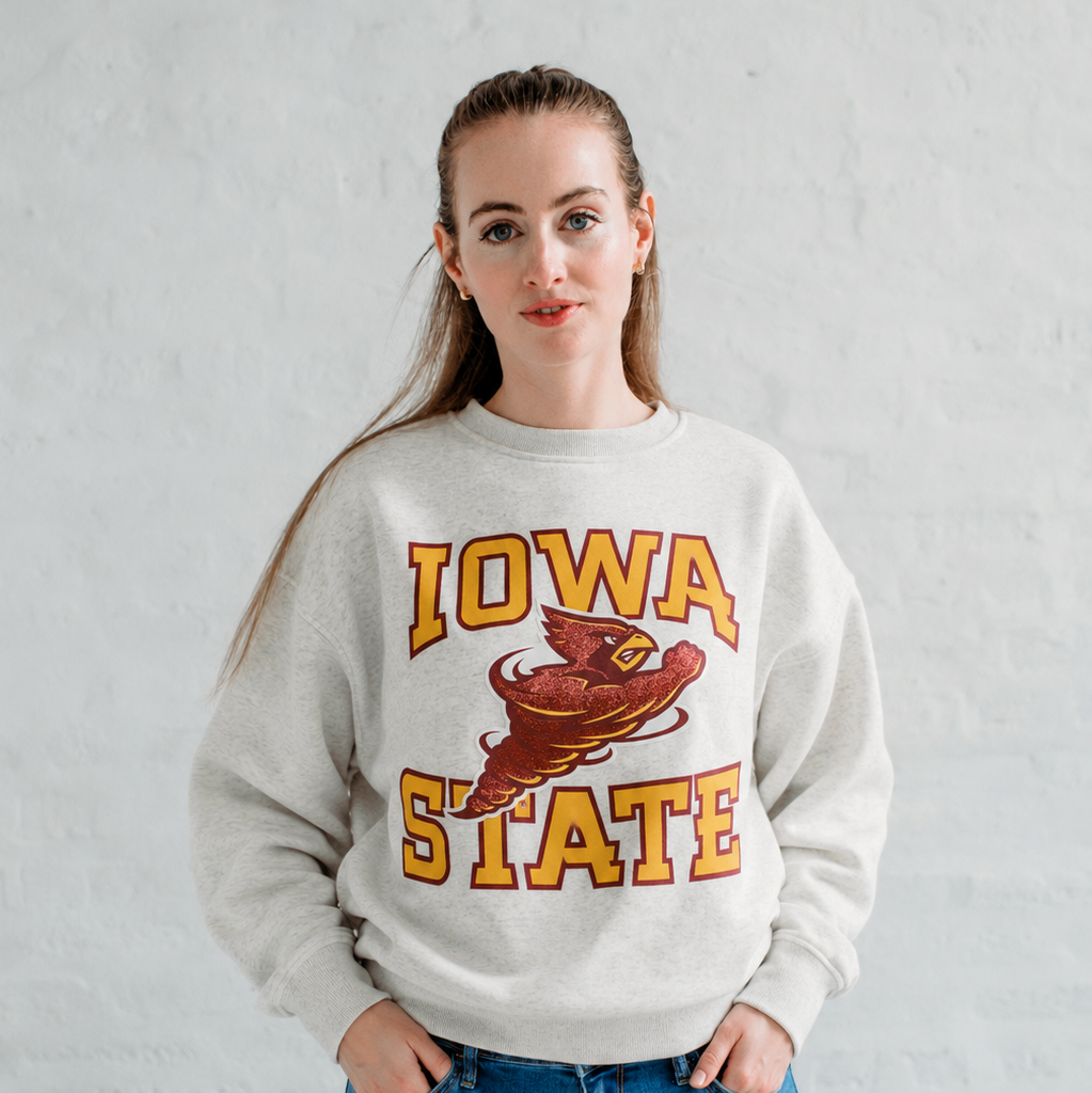 Model wearing the Iowa State Football Slouchy Drop Shoulder Crewneck in oatmeal heather, featuring vintage Iowa State lettering and the Cy mascot graphic across the front.