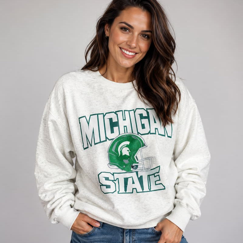 Model wearing the Michigan State Spartans Football Slouchy Drop Shoulder Crewneck in oatmeal heather, featuring Michigan State lettering and a green Spartan football helmet graphic.