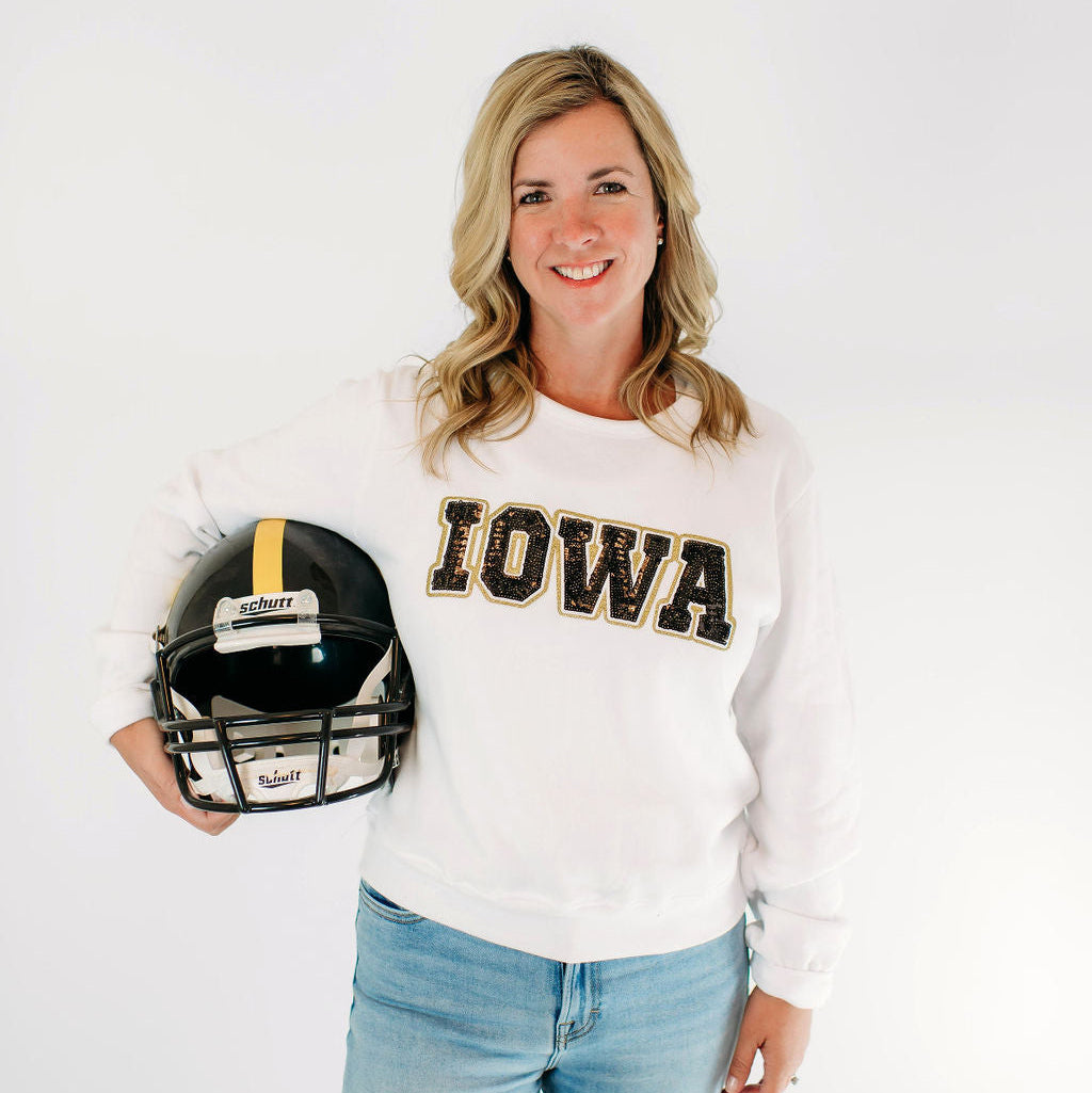 Model wearing the Iowa Hawkeyes Women's Sequin Crewneck in white, featuring black sequin IOWA lettering outlined in gold across the chest, while holding a black football helmet.