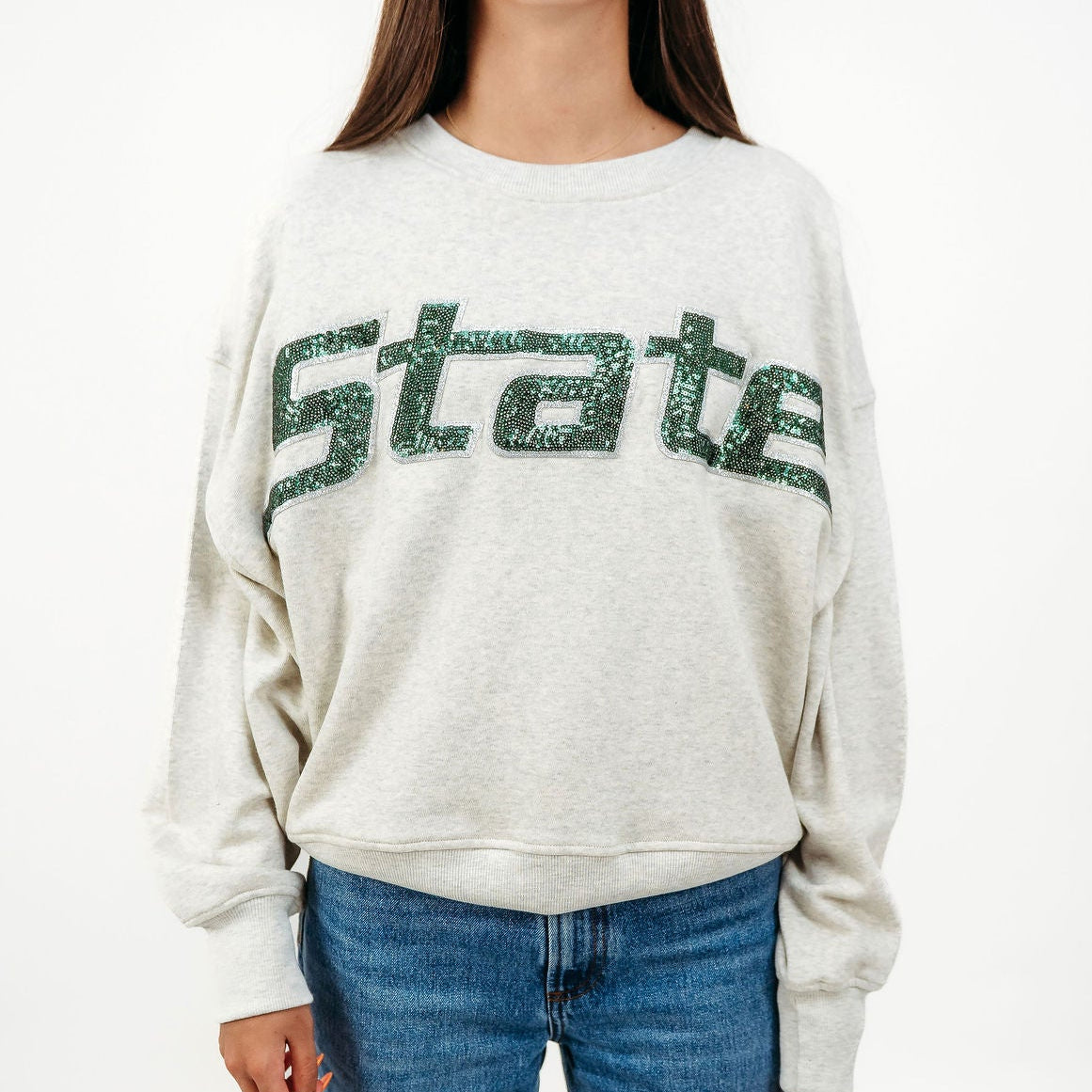 A woman wearing a light heather gray crewneck sweatshirt with 'State' written across the chest in a glittery green font, paired with blue jeans, against a plain white background.