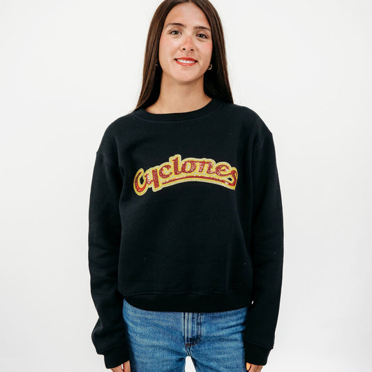 A young woman with long brown hair stands against a white studio background wearing a black crewneck sweatshirt with 'Cyclones' written across the chest in a yellow and orange retro font, paired with blue jeans.