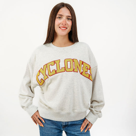 A woman modeling a light heather gray crewneck sweatshirt with 'CYCLONE' printed across the chest in yellow and orange varsity lettering, paired with blue jeans against a plain white studio background.