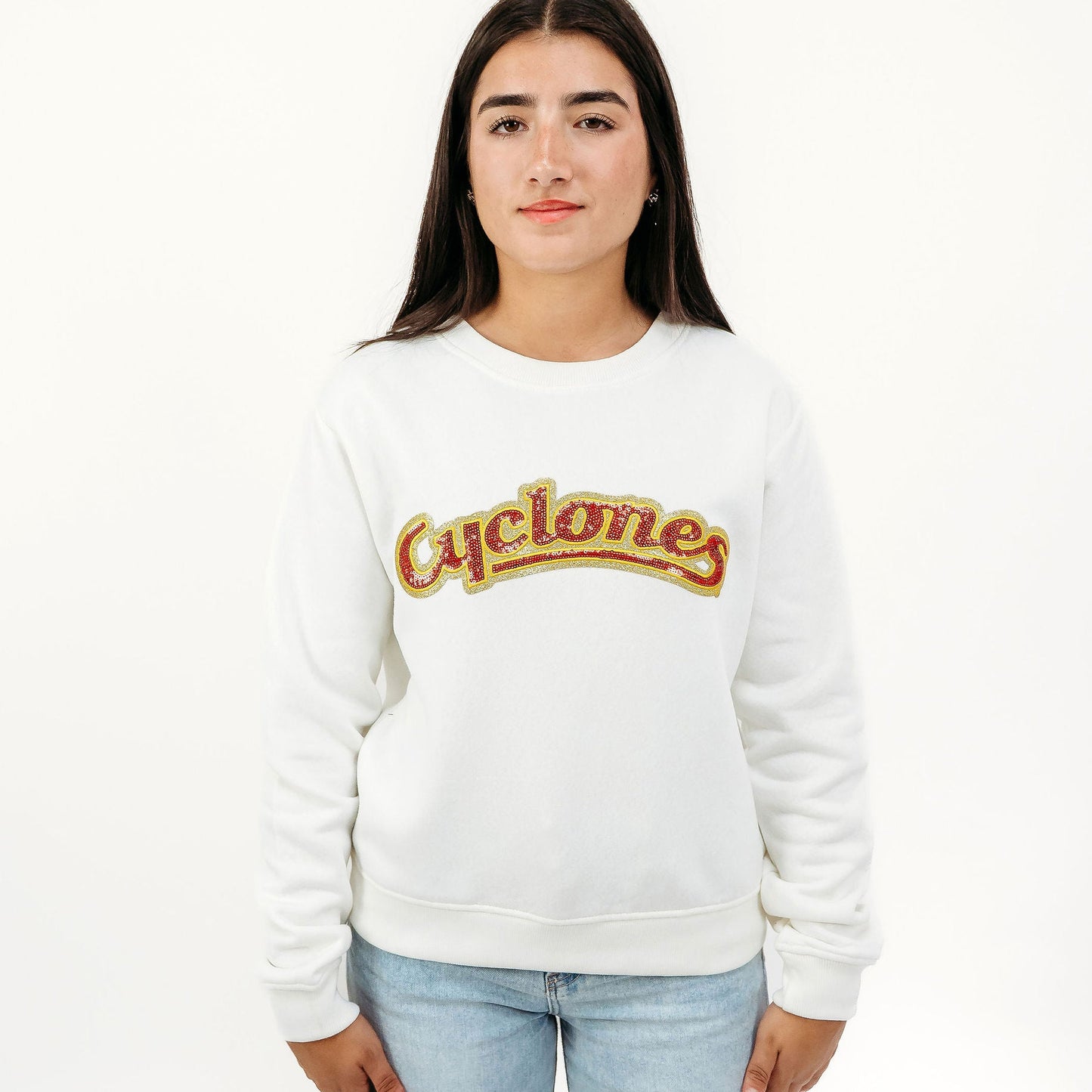 A young woman modeling a white crewneck sweatshirt with a retro-style 'Cyclones' logo across the chest in red and gold, paired with light blue jeans, against a plain white studio background.