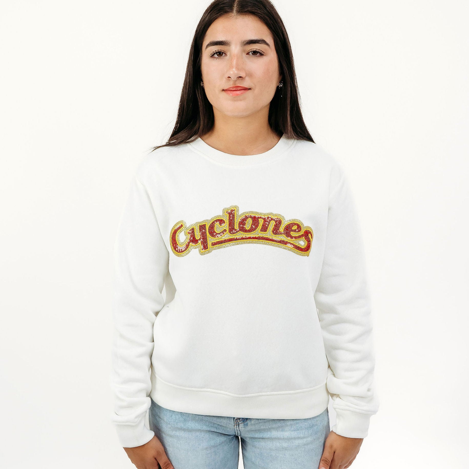 A young woman modeling a white crewneck sweatshirt with a retro-style 'Cyclones' logo across the chest in red and gold, paired with light blue jeans, against a plain white studio background.